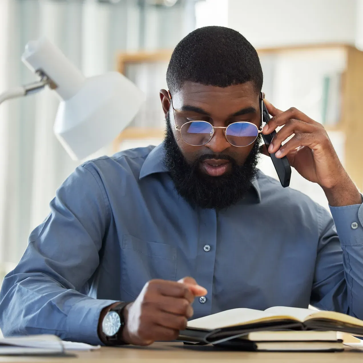 A criminal justice professional in a blue dress shirt and glasses reads a legal book while speaking on the phone, with a scale of justice and a desk lamp in the background.