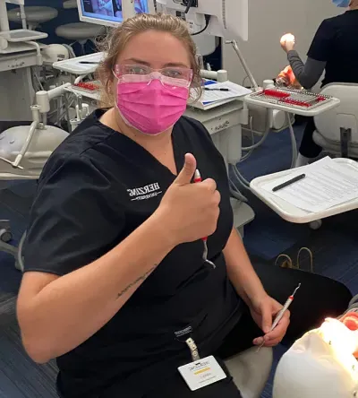 A Herzing dental hygiene student giving a thumbs up while performing a dental procedure. The student is wearing a pink mask and protective glasses, with a dental monitor showing images in the background. A patient is seated in the dental chair, receiving treatment.